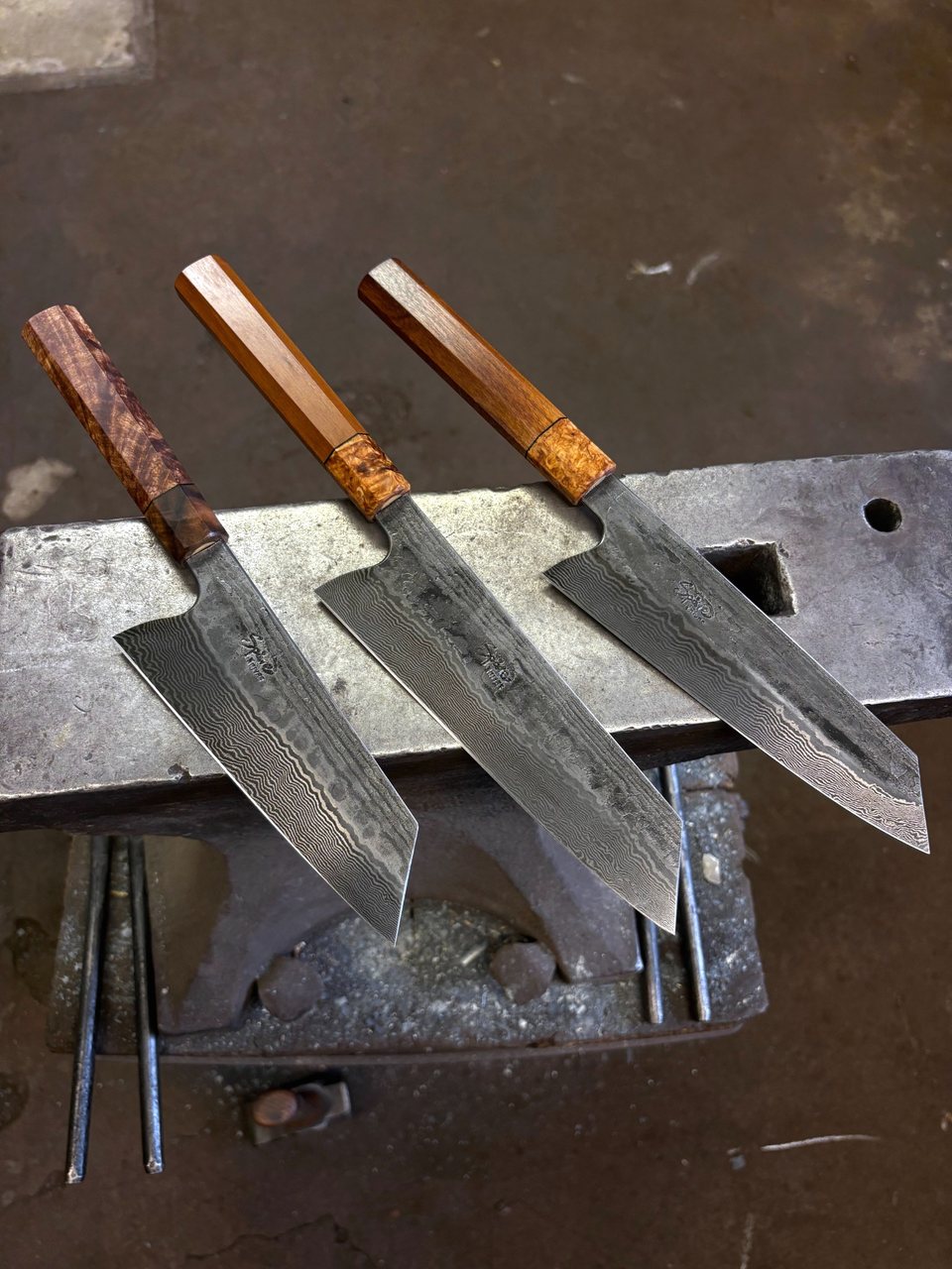 Three finished chef knives with damascus patterned blades and octagonal wooden handles lie on an anvil