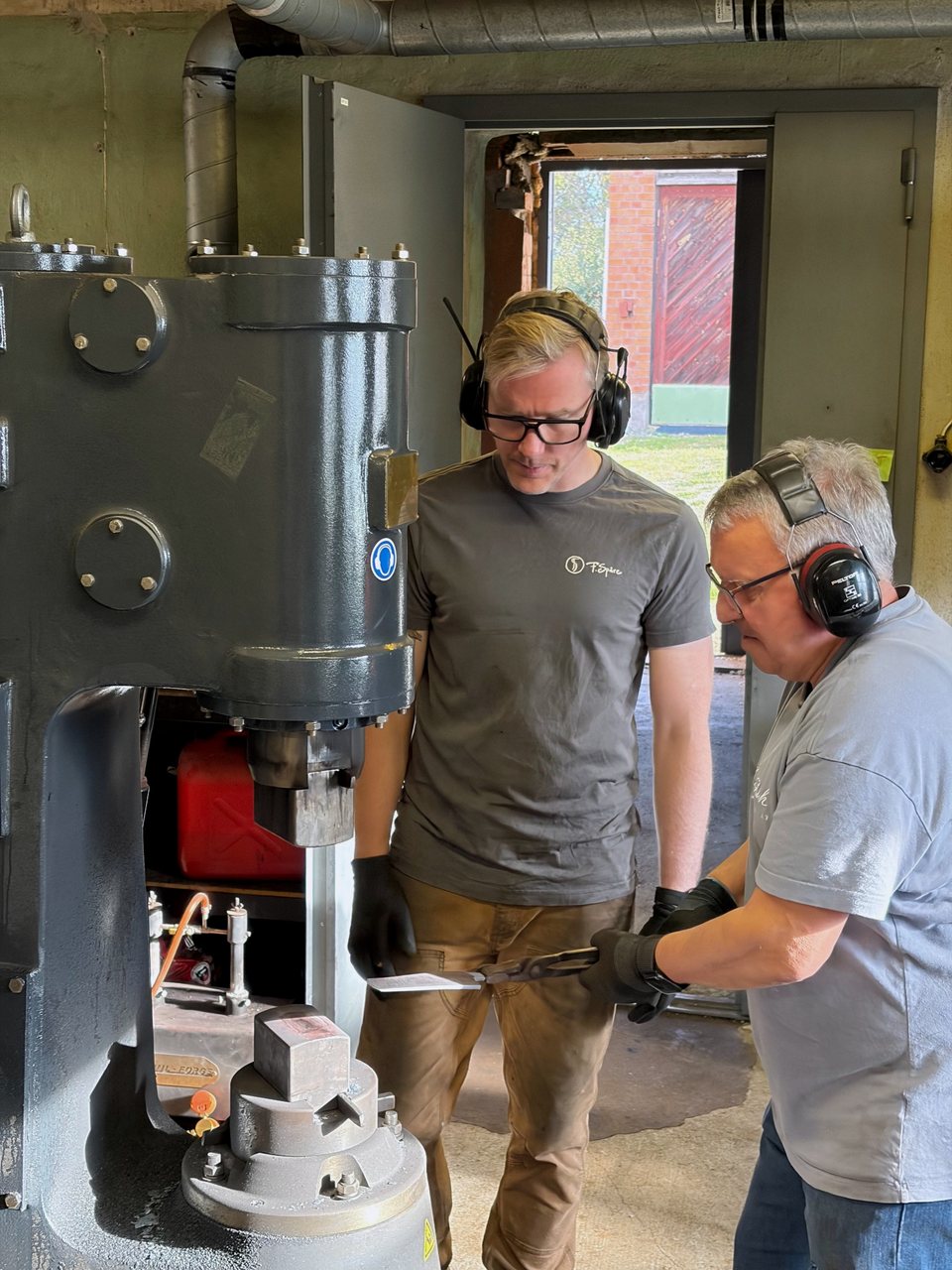 An instructor and a class participant in ear defenders hold a workpiece with tongs at the big power hammer in the workshop